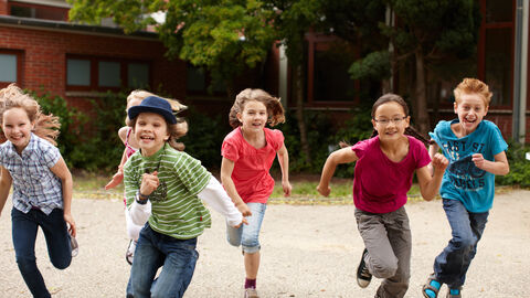 Bewegt aufwachsen Sechs Kinder im Grundschulalter rennen mit Elan und Begeisterung auf einem Schulhof auf die Kamera zu. Im Hintergrund ist der Schulhof mit Bäumen und einem Gebäude zu sehen.