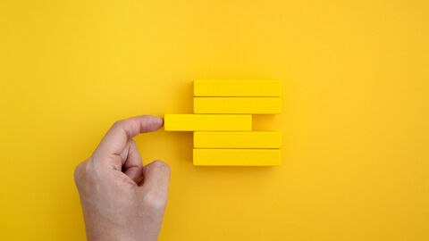 Chancengleichheit Vor einem gelben Hintergrund liegen 5 gelbe Holzbausteine übereinander. Der mittlere Baustein ist etwas nach links verlagert. Links von den Bausteinen ist eine Hand zu sehen, die mit dem Zeigefinger den herausschauenden Baustein wieder gerade zurückschieben möchte.