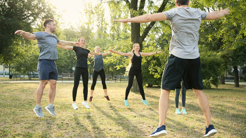 Bewegungskoordinatoren In einem Park sind sechs springende junge Erwachsene in Sportkleidung zu sehen. Beine und Arme sind hierbei weit auseinandergestreckt. Vorne rechts ist von hinten der vermutliche Übungsleiter zu sehen, alle weiteren Erwachsenen schauen ihn hierbei lächelnd an.