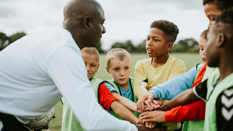 Integration und Sport Auf einem Fußballfeld im Freien sind im Vordergrund ein Coach und fünf Jungen mit und ohne Migrationshintergrund im Sporttrikot abgebildet, die alle ihre Hände aufeinander gestalpelt halten. Hierbei schauen sie sich alle in die Augen.