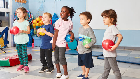 Bewegungsförderung in der Kita In einem Sportraum mit Sportmatten und weiteren Sportaccessoires, stehen im Vordergrund fünf Kleinkinder mit und ohne Migrationshintergrund nebeneinander. Alle Kinder halten jeweils einen Ball in unterschiedlichen Farben in den Händen und sind sportlich gekleidet.