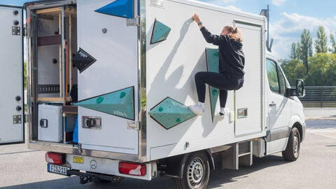 Vielfältiges Bewegungsprogramm auf dem Hafenparkgelände zum Mitmachen: Sportmobil Adventure Cube (Sportjugend Hessen) Vielfältiges Bewegungsprogramm auf dem Hafenparkgelände zum Mitmachen: Sportmobil Adventure Cube (Sportjugend Hessen). Hier klettert eine junge Frau an der Wand eines Transporters der als Boulderwand dient.