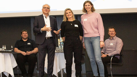 Der Hessische Minister für Wirtschaft, Energie, Verkehr und Wohnen Tarek Al-Wazir steht auf einer Bühne und gratuliert Yvonne Ramus (rechts) und Béatrice Frank (mitte) von der HAGE zur Zertifizierung “Fahrradfreundlicher Arbeitgeber”.