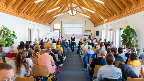 Hauptraum der Fachtagung zu Beginn der Veranstaltung. Der Saal ist mit Menschen gefüllt, die ihre Aufmerksamkeit nach vorne zur Moderatorin richten.