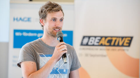 Fachforum 2: bewegt bleiben in der Kommune, Vortrag Marco Mattes: Sport im Park Marco Mattes mit Mikro in der Hand, im Hintergrund sind Roll-Ups zu sehen.