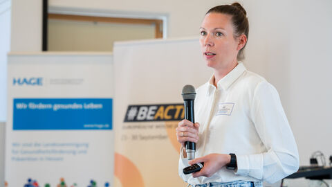 Fachforum 2: bewegt bleiben in der Kommune, Vortrag Miriam Roth: Städtebauförderung Miriam Roth während Ihres Vortrages mit Mikro und Pointer in der Hand, im Hintergrund sind Roll-Ups zu sehen