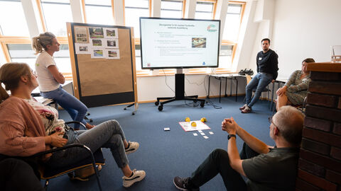Fachforum 1: bewegt aufwachsen in der Kommune, Gruppenarbeit Man sieht insgesamt fünf Personen, die sich um einen TV-Bildschirm und eine Pinnwand im Kreis platziert haben. Zwei Personen sitzen auf Stühlen, eine auf dem Boden und zwei lehnen an Tischen. Auf dem Boden liegen und an der Pinwand hängen Arbeitsmaterialien. Der Bildschirm zeigt die Folie einer Präsentation.