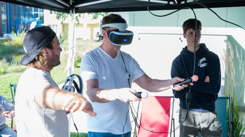 Bewegter Ausklang: Virtual Reality Mann trägt eine Virtual-Reality-Brille und hat zwei Controler in der Hand. Er wird dabei von einem Übungsleiter angeleitet.