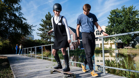 Bewegter Ausklang: E-Longboard Frau fährt auf elektronischem Longboard über einen Steg im Außenbereich. Sie trägt einen Helm. Ein Übungsleiter läuft neben ihr her zur Sicherheit.