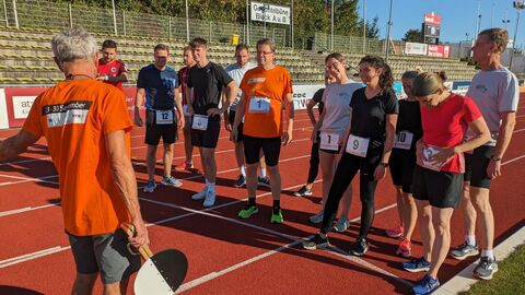 Es sein einige Menschen zu sehen, die in einer Reihe nebeneinander stehen. Sie tragen Sportklamotten und stehen auf der roten Laufbahn eines Sportplatzes. Alle haben Schilder mit Startnummern an ihren Shirts. Vor der Reihe steht ein Herr mit Startklappe in der Hand, diese ist nach unten gesengt. Er erklärt den Teilnehmern die Aufgabe