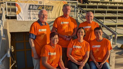 Es sind 6 Personen zu sehen. In der ersten Reihe sitzen drei Frauen auf einer Bank. Hinter ihnen in der zweiten Reihe stehen drei Herren. Im Hintergrund ist eine Tribüne zu sehen und ein Banner von BeActive. Alle tragen orangene T-Shirts der Bewegungskampagne. 