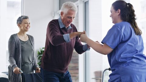 Bewegungsförderung in Pflegeeinrichtungen Pflegerin hilft älterem Herr aus dem Rollstuhl. Sie steht vor ihm und hält beide Hände Der Herr mit grauen Haaren steht bereits fast vollständig und lächelt. Der Rollstuhl wird von einer dritten weiblichen Person festgehalten.  Auch sie lacht.