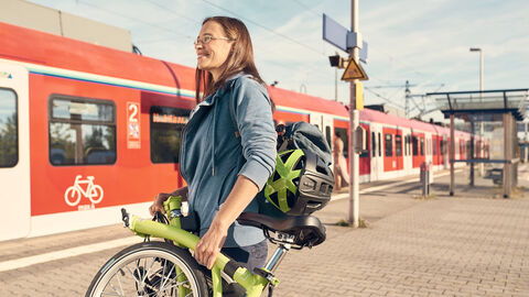 8. Nahmobilitätskongress Hessen Bei schönem Wetter ist eine Frau mit eingeklappten Fahrrad auf einem Bahngleis zu sehen. Im Hintergrund steht ein eingefahrener roter Regionalzug.