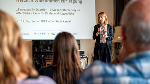 Die Gesundheits- und Jugenddezernentin Nicole Maisch begrüßt die Teilnehmenden. Im Hintergrund ist eine Leinwand mit dem Titel der Tagung zu sehen. Im Vordergrund sind Teilnehmende von hinten zu sehen (verschwommen).