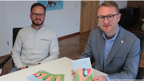 Zwei Personen sitzen an einem Tisch und Präsentieren ein Heft 