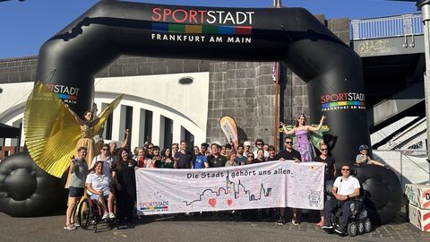 Gruppenbild des zweiten Inklusionslaufs in Frankfurt  Gruppe von Menschen, die ein Banner hochhalten unter einem aufblasbaren Torbogen des Sportamts.