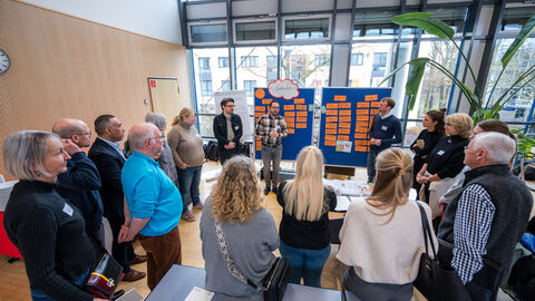 Menschen stehen vor Pinnwand und hören Redner zu 