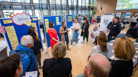 Menschen stehen um Pinnwand und hören Sprecherin zu 