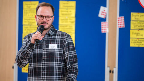 Mann mit Mikrofon in der Hand vor Pinnwand