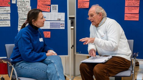 Zwei Personen sitzen vor einer Pinwand auf Stühlen und unterhalten sich angeregt.