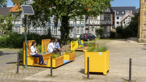 Das Bild zeigt eine urbane Sitzgelegenheit im Freien mit integrierten Pflanzenkästen in einer gepflasterten Platzanlage. Die Sitzbänke aus Holz haben gelbe Akzente und bieten Platz für mehrere Personen, von denen drei dort sitzen und sich unterhalten oder auf ihr Smartphone schauen. Im Hintergrund sind Fachwerkhäuser, ein Baum sowie ein Auto zu sehen. Ein solarbetriebenes Lichtmast ist ebenfalls erkennbar.