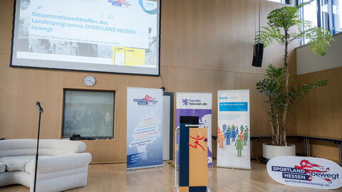 Auf dem Bild ist eine Projektionsleinwand mit Willkommensfolie zu sehen, und darunter drei Roll-Ups und ein Rednerpult.