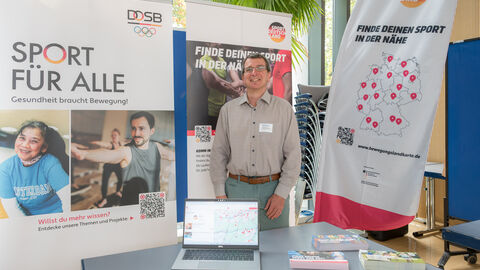 Ein Aussteller steht an seinem Stand, hinter ihm ist ein Roll-Up und eine Pinnwand zu sehen.