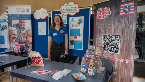 Eine Ausstellerin steht an ihrem Stand, hinter ihr ist ein Roll-Up und eine Pinnwand zu sehen.
