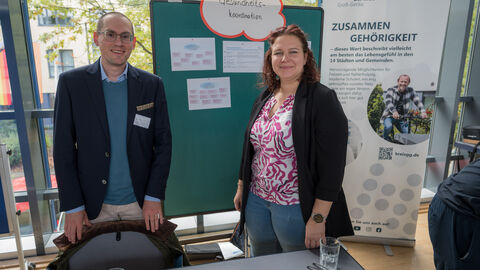 Zwei Aussteller stehen an ihrem Stand, hinter ihnen ist ein Roll-Up und eine Pinnwand zu sehen.